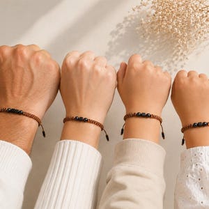 May include: A set of four matching bracelets featuring a brown woven cord and three central black beads, including two spherical beads flanking a single square bead. The bracelets are adjustable with dangling cord ends and a knotted design.