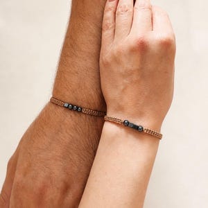 Puede incluir: Dos pulseras de cordón trenzado color canela con detalles metálicos oscuros. Una pulsera muestra tres pequeñas cuentas esféricas y una cuadrada, mientras que la otra presenta dos cuentas esféricas flanqueando una cuenta cuadrada central.