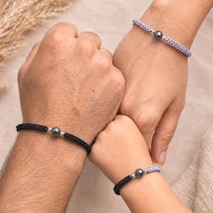 May include: Three braided cord bracelets featuring a central faceted dark gray bead flanked by two small silver-toned metallic beads. One bracelet is black, one is light purple, and the third is a combination of black and light purple cord.