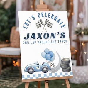 May include: A white rectangular sign featuring the text LET'S CELEBRATE JAXON'S 2ND LAP AROUND THE TRACK, two crossed checkered flags, a light blue race car with the number 2, a cluster of blue balloons, and a stack of three black tires.