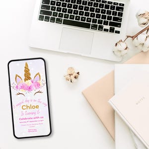 May include: A smartphone screen shows a unicorn-themed birthday invitation. The design includes a gold horn, pink flowers, and the text "Chloe is turning 6." A laptop, notebooks, and cotton stems are arranged on a white surface.