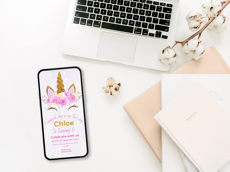May include: A smartphone screen shows a unicorn-themed birthday invitation. The design includes a gold horn, pink flowers, and the text "Chloe is turning 6." A laptop, notebooks, and cotton stems are arranged on a white surface.