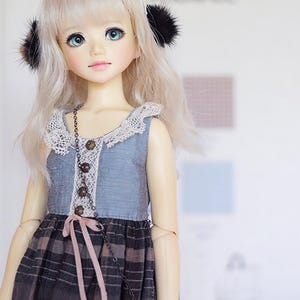 May include: A ball-jointed doll with long blonde hair, teal eyes, and black earmuffs. The doll wears a blue sleeveless dress with a white lace collar, three small brown buttons, and a dark brown and black plaid skirt with a pink ribbon tie.