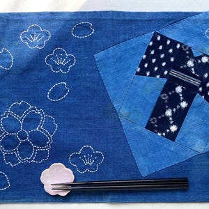 May include: A blue placemat with white embroidered floral designs and a patchwork pattern. A pink ceramic chopstick rest and black chopsticks are on the placemat. The placemat is made of fabric.