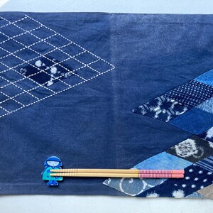 May include: A navy blue placemat with a geometric design. The placemat features a diamond pattern stitched with white thread and a patchwork design in shades of blue. A small blue figurine and a pair of chopsticks are placed on the placemat.