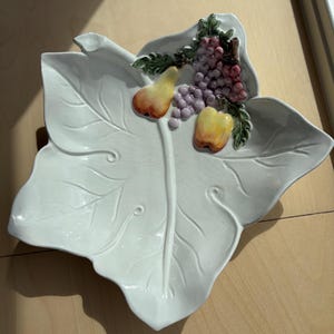 Op de afbeelding: Een witte keramische schaal in de vorm van een groot blad met een glanzende afwerking en een craquelé textuur. De schaal is voorzien van een verhoogd, driedimensionaal decor van een gele peer, een rode appel en paarse druiven.