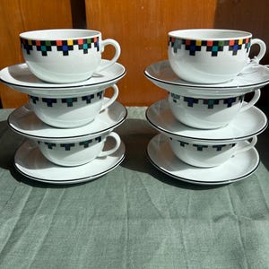 Puede incluir: Seis tazas de té y platillos de cerámica blanca dispuestos en dos pilas de tres. Cada taza presenta un patrón geométrico repetitivo alrededor del borde con cuadrados negros, rojos, amarillos y turquesas. Los platillos tienen una línea negra fina.