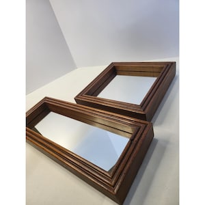 May include: Two mirrors with thick, dark brown wooden frames. One mirror is square-shaped, and the other is rectangular. Both frames feature a stepped, layered design with visible wood grain texture, surrounding clear, reflective glass surfaces.