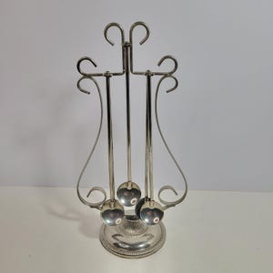 May include: A silver-toned metal stand featuring a lyre-shaped frame with decorative scrollwork, designed to hold three small spoons. The stand rests on a circular, ribbed base, and the three spoons hang vertically from the top hooks of the frame.