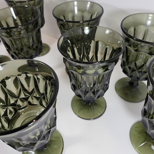 Può includere: Una collezione di calici verde oliva in stile vintage. Ogni bicchiere presenta un motivo a taglio diamante sulla coppa e una base arrotondata. I bicchieri sono disposti su una superficie bianca, mostrando il loro design e colore unici.