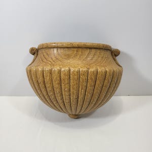 May include: A light brown, semi-circular wall planter featuring a prominent wood grain texture and vertical fluted detailing. The vessel has a wide, open top rim with small, curled decorative handles on each side and a single point at the base.
