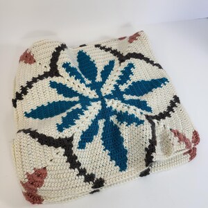 May include: A crocheted square blanket featuring a central teal floral design, surrounded by dark gray and pink accents on an off-white background. The blanket is folded, showcasing the intricate stitch work and color palette.