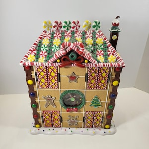 May include: A decorative gingerbread house advent calendar featuring a brown textured facade, a roof adorned with colorful candy-like decorations, a central clock, and hinged doors. The structure includes a chimney and a white base with sprinkles.