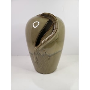 May include: A glossy olive green ceramic vase with a smooth, rounded body and a distinctive vertical slit opening that curves along the upper section. The surface features a subtle, embossed leaf pattern near the base and a reflective glaze finish.