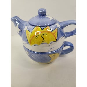 May include: A ceramic tea for one set featuring a blue background with white stars and clouds. The teapot lid and body display a yellow sun with a face, while the matching cup below features a yellow crescent moon with a closed eye and smile.