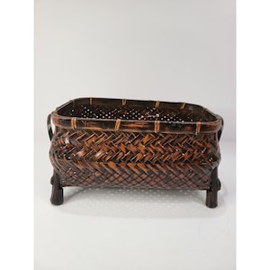 May include: A rectangular woven basket featuring a dark brown and black finish with a herringbone pattern. The basket has a reinforced rim, two side handles, and stands on four short, sturdy legs, showcasing a detailed, interlaced construction.