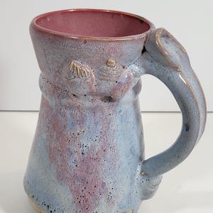 May include: A handmade ceramic mug with a unique design. The mug features a light blue and purple glaze with a textured surface. The handle is a light blue color. The interior of the mug is a dark pink color. The mug has decorative elements near the handle.