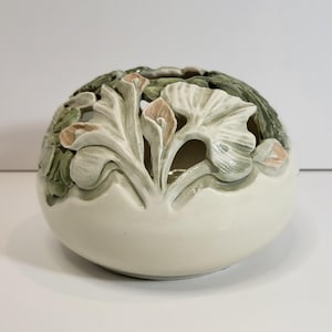 Puede incluir: Un portavelas esférico de cerámica con diseño floral. La base blanquecina está adornada con calas y hojas talladas en tonos verdes y melocotón. La parte superior tiene secciones recortadas para que pase la luz.