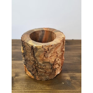 Puede incluir: Un recipiente cilíndrico de madera con un exterior de corteza natural y rugosa en tonos marrones y bronceados. El borde superior es liso y circular, revelando la veta de la madera. El objeto tiene un centro hueco y una estructura robusta.