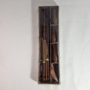 May include: A rectangular brown box containing a set of dark brown wooden chopsticks and three carved wooden fish-shaped chopstick rests. A decorative brown leaf-shaped accent is attached to the chopsticks. The word Maui is printed on the box.