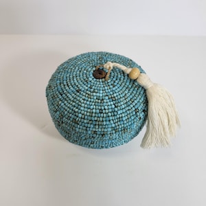 May include: A round, turquoise beaded box with a wooden button closure and a white tassel. The box is covered in small, light blue beads, creating a textured surface. The tassel is attached with a cream-colored cord.