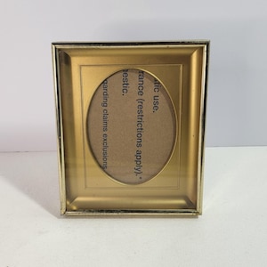 May include: A rectangular gold-toned picture frame featuring an oval-shaped central opening. The frame includes a cardboard insert with printed text that reads: stic use. rance (restrictions apply).* garding claims exclusions estic.