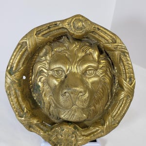 Pode incluir: Um aldravão de porta circular em latão com uma cabeça de leão detalhada no centro. O leão é rodeado por uma moldura decorativa com um padrão trançado. O metal tem um acabamento dourado desgastado com pátina escura nas reentrâncias.