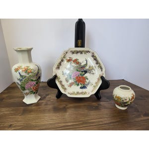 May include: A collection of three cream-colored ceramic pieces featuring colorful peacock and floral motifs. The set includes a tall hexagonal vase, a wide octagonal bowl displayed on a black stand, and a small, round, bulbous vase.