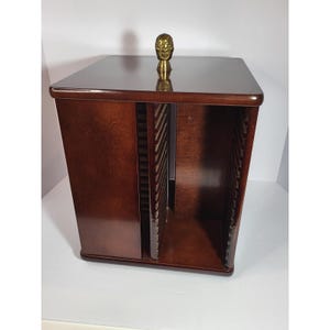 May include: A square wooden media storage tower with a dark brown finish and a central rotating mechanism. The top features a small, ornate brass-colored finial. The unit contains multiple vertical slots designed for organizing discs.