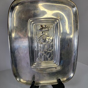 May include: A rectangular silver-toned metal tray with rounded corners. The centre features a raised, embossed design of a figure carrying a basket on their head, surrounded by stylised foliage and cacti. The surface has a polished, reflective finish.