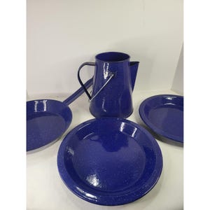 Puede incluir: Un juego de utensilios de esmalte azul oscuro con un patrón de motas blancas. La colección incluye una cafetera alta con asa de metal, una sartén con mango largo y dos platos redondos. Cada pieza tiene un acabado liso, brillante y uniforme.