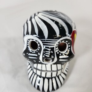 May include: A ceramic skull figurine featuring a glossy black base with hand-painted white decorative patterns. The design includes circular eye sockets, a small nasal cavity, and a row of teeth, accented by a small red detail on the side.