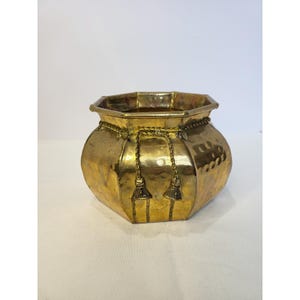 Puede incluir: Una maceta de latón pulido con forma octogonal y textura martillada. El recipiente presenta un detalle decorativo de cuerda de latón que rodea el borde superior y cuelga por la parte frontal en dos hilos distintos con borlas pequeñas.