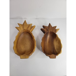 May include: Two carved wooden bowls shaped like pineapples. Each bowl features a hollowed-out centre with a natural wood grain pattern, a pointed top section representing leaves, and a rounded base. The wood displays warm brown tones.