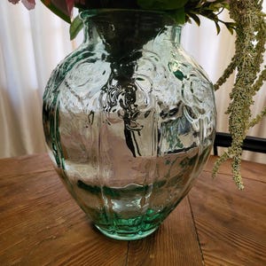 Puede incluir: Un jarrón de vidrio transparente con un tinte verde, con una superficie texturizada con patrones florales. El jarrón está lleno de flores rosas y vegetación, sobre una mesa de madera. El jarrón tiene un cuerpo ancho y redondeado y un cuello estrecho.