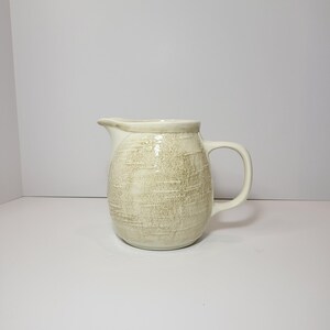 May include: A cream-colored ceramic pitcher with a textured surface. The pitcher has a handle and a spout for pouring. The design is simple and rustic, suitable for serving beverages or as a decorative item.