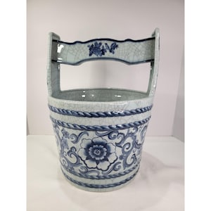 Könnte beinhalten: Ein Keramikeimer mit hellblauer Craquelé-Glasur, verziert mit einem dunkelblauen Blumenmuster und einer seilartigen Bordüre. Der Eimer verfügt über einen hohen, gewölbten Henkel mit einem kleinen blauen Blumenmotiv in der Mitte.