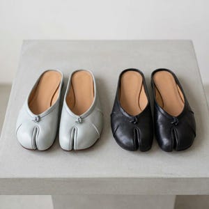 May include: Two pairs of leather slip-on shoes. One pair is light blue, the other is black. Both have a split-toe design and a small knot detail at the top. The shoes have a tan-colored insole.