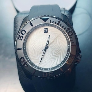 May include: A wristwatch with a black silicone strap and a dark metallic bezel featuring engraved numbers. The watch face is white with a subtle, wavy, rippled texture, black hands, and a small black logo at the twelve o'clock position.