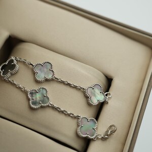 K&ouml;nnte beinhalten: Ein silbernes Armband mit Kleeblatt-Anh&auml;ngern, die jeweils mit einer Perlmutt-Einlage versehen sind. Das Armband wird in einer beigen Schmuckschatulle pr&auml;sentiert. Die Anh&auml;nger sind mit einem Perlenmuster umrandet.