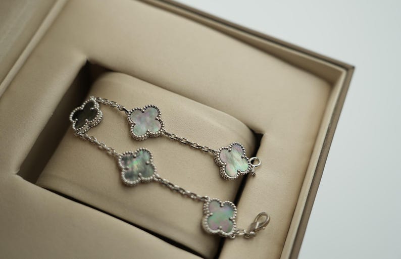 K&ouml;nnte beinhalten: Ein silbernes Armband mit Kleeblatt-Anh&auml;ngern, die jeweils mit einer Perlmutt-Einlage versehen sind. Das Armband wird in einer beigen Schmuckschatulle pr&auml;sentiert. Die Anh&auml;nger sind mit einem Perlenmuster umrandet.