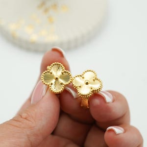 K&ouml;nnte beinhalten: Ein Paar goldfarbene Ohrringe in Kleeblattform. Jeder Ohrring hat ein vierbl&auml;ttriges Kleeblatt-Design mit strukturierter Oberfl&auml;che und einem kleinen zentralen Detail. Die Ohrringe werden in einer Hand gehalten.