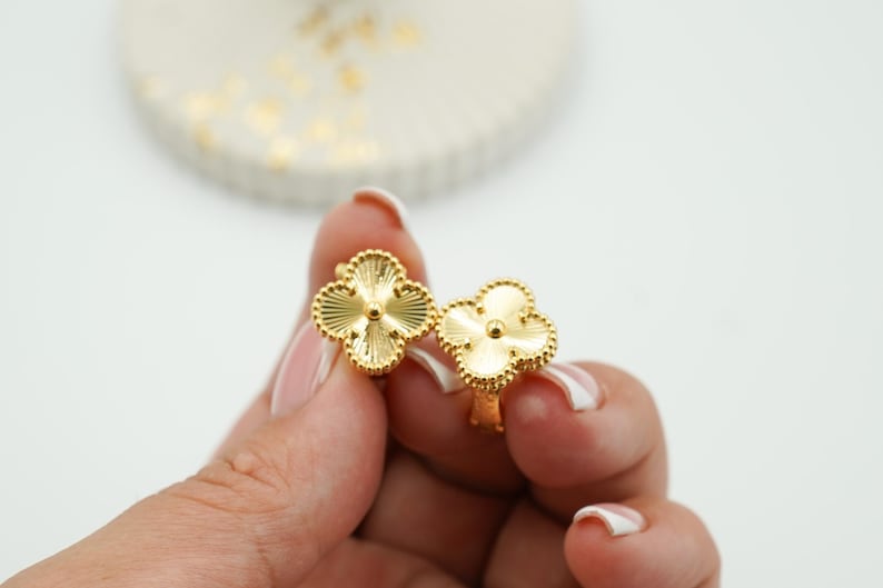 K&ouml;nnte beinhalten: Ein Paar goldfarbene Ohrringe in Kleeblattform. Jeder Ohrring hat ein vierbl&auml;ttriges Kleeblatt-Design mit strukturierter Oberfl&auml;che und einem kleinen zentralen Detail. Die Ohrringe werden in einer Hand gehalten.