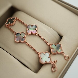 K&ouml;nnte beinhalten: Ein ros&eacute;goldenes Armband mit Kleeblatt-Anh&auml;ngern, die jeweils mit einer Perlmutt-Einlage versehen sind. Das Armband wird in einer beigen Schmuckschatulle pr&auml;sentiert. Die Kleeblatt-Anh&auml;nger haben einen gewellten Rand.