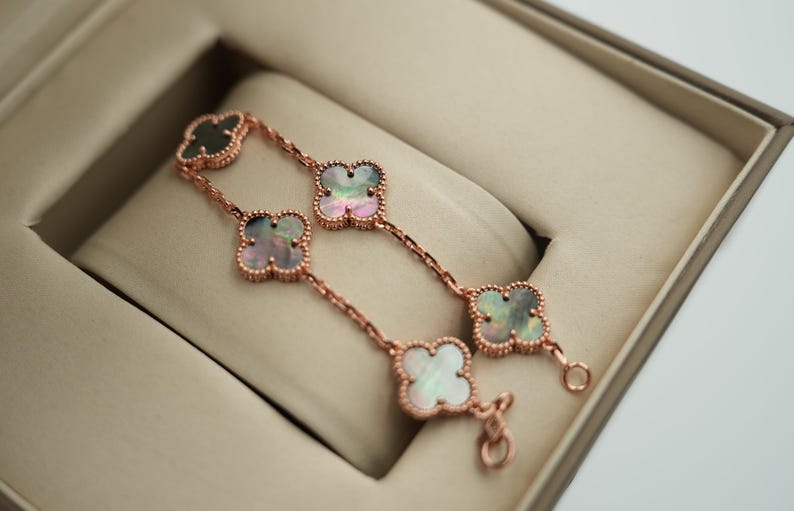 K&ouml;nnte beinhalten: Ein ros&eacute;goldenes Armband mit Kleeblatt-Anh&auml;ngern, die jeweils mit einer Perlmutt-Einlage versehen sind. Das Armband wird in einer beigen Schmuckschatulle pr&auml;sentiert. Die Kleeblatt-Anh&auml;nger haben einen gewellten Rand.