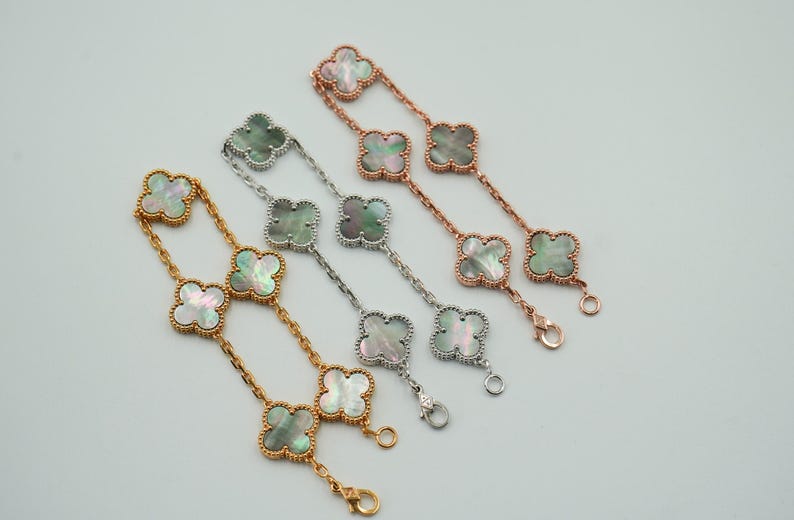 K&ouml;nnte beinhalten: Vier Armb&auml;nder mit Kleeblatt-Anh&auml;ngern. Die Armb&auml;nder sind in Gold-, Silber- und Ros&eacute;goldt&ouml;nen gehalten. Jeder Kleeblatt-Anh&auml;nger hat einen Perlmutt-Mittelteil. Die Armb&auml;nder werden auf einer wei&szlig;en Oberfl&auml;che pr&auml;sentiert.