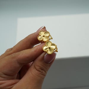 K&ouml;nnte beinhalten: Ein Paar goldfarbene Ohrringe in Kleeblattform. Jeder Ohrring hat ein vierbl&auml;ttriges Kleeblatt-Design mit strukturierter Oberfl&auml;che und einem zentralen Akzent. Die Ohrringe werden in einer Hand vor wei&szlig;em Hintergrund gehalten.