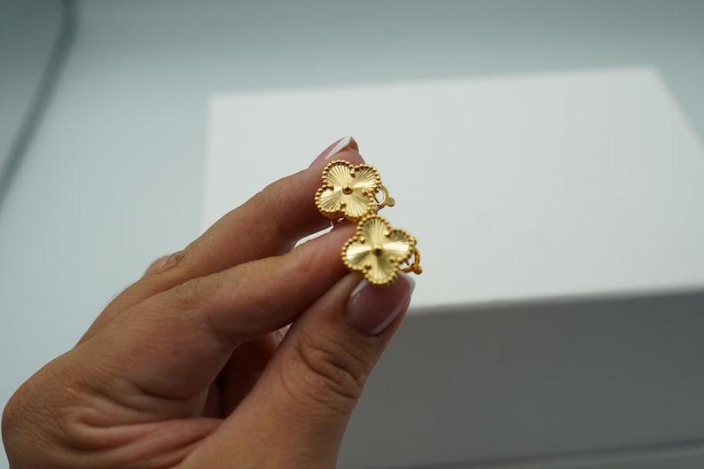 K&ouml;nnte beinhalten: Ein Paar goldfarbene Ohrringe in Kleeblattform. Jeder Ohrring hat ein vierbl&auml;ttriges Kleeblatt-Design mit strukturierter Oberfl&auml;che und einem zentralen Akzent. Die Ohrringe werden in einer Hand vor wei&szlig;em Hintergrund gehalten.