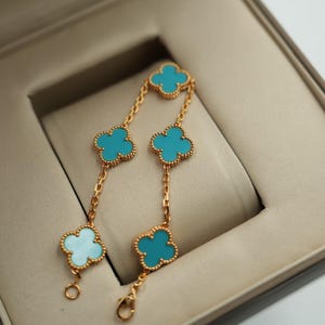Puede incluir: Pulsera dorada con cinco colgantes en forma de tr&eacute;bol de color turquesa. La pulsera se muestra dentro de una caja de joyer&iacute;a beige. Los colgantes de tr&eacute;bol est&aacute;n conectados por delicadas cadenas doradas.