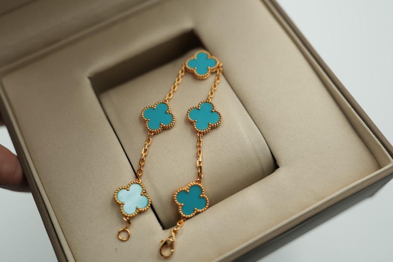 Puede incluir: Pulsera dorada con cinco colgantes en forma de tr&eacute;bol de color turquesa. La pulsera se muestra dentro de una caja de joyer&iacute;a beige. Los colgantes de tr&eacute;bol est&aacute;n conectados por delicadas cadenas doradas.